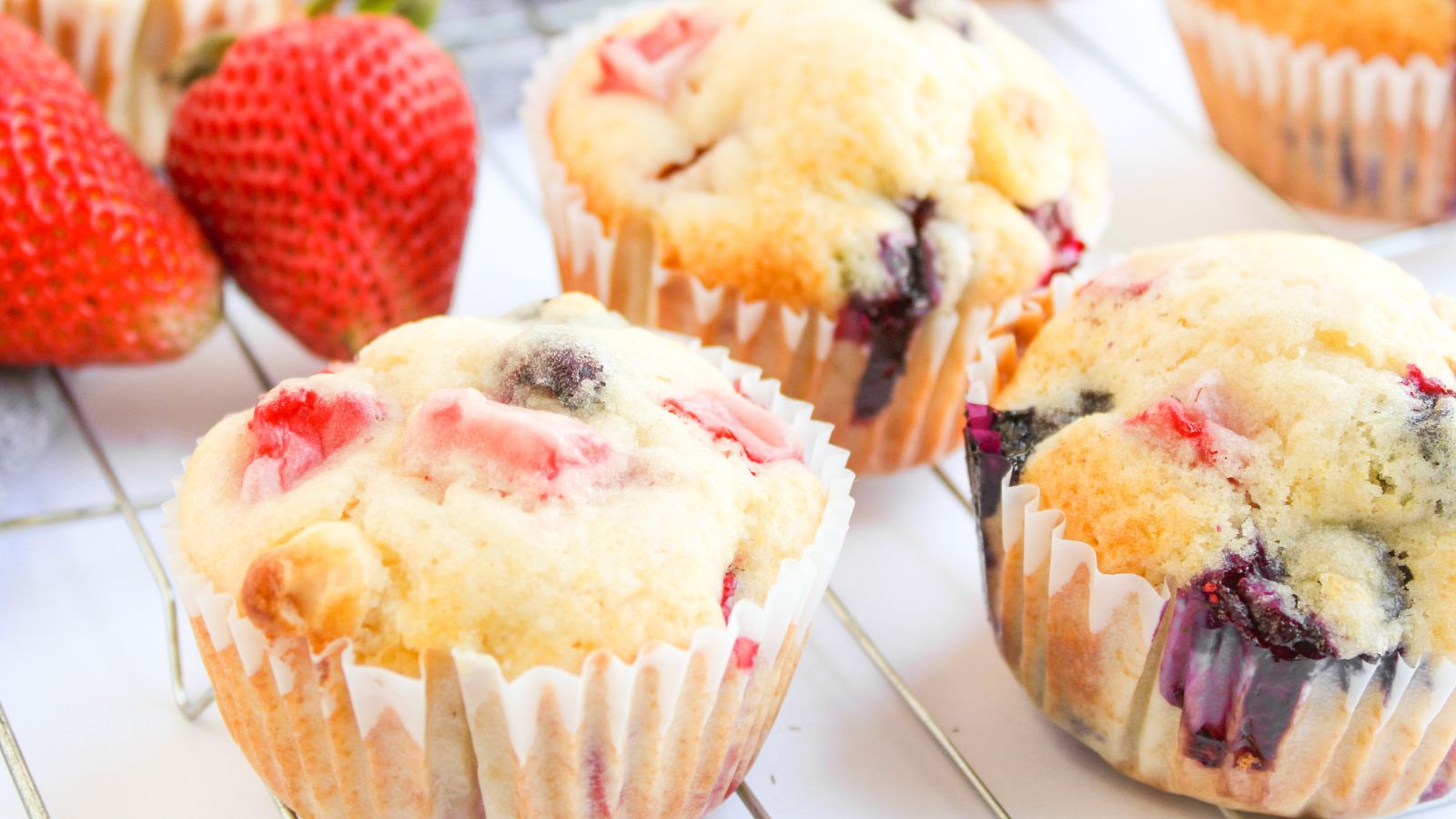 Three berry muffins with visible strawberry and blueberry pieces sit on a cooling rack next to whole strawberries.