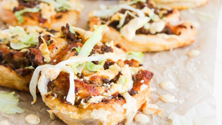 Close-up of puff pastry rounds topped with ground beef, shredded lettuce, sesame seeds, sauce, and melted cheese on parchment paper.
