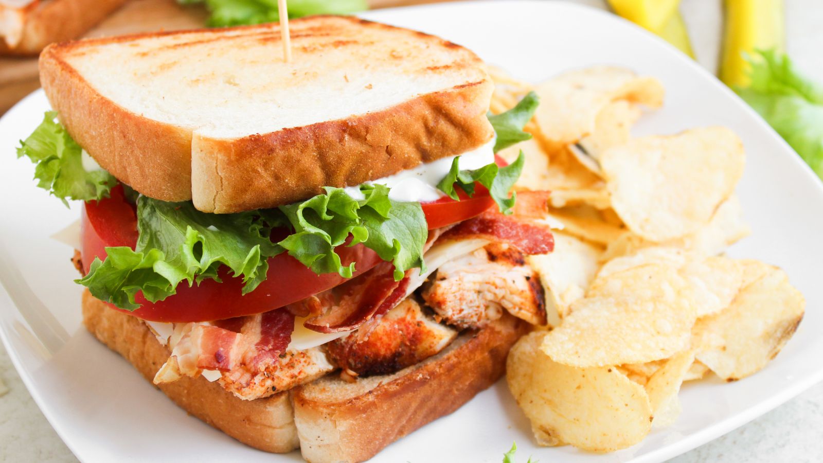A grilled chicken sandwich with lettuce, tomato, bacon, and mayonnaise on toasted bread, served with potato chips on a white plate.