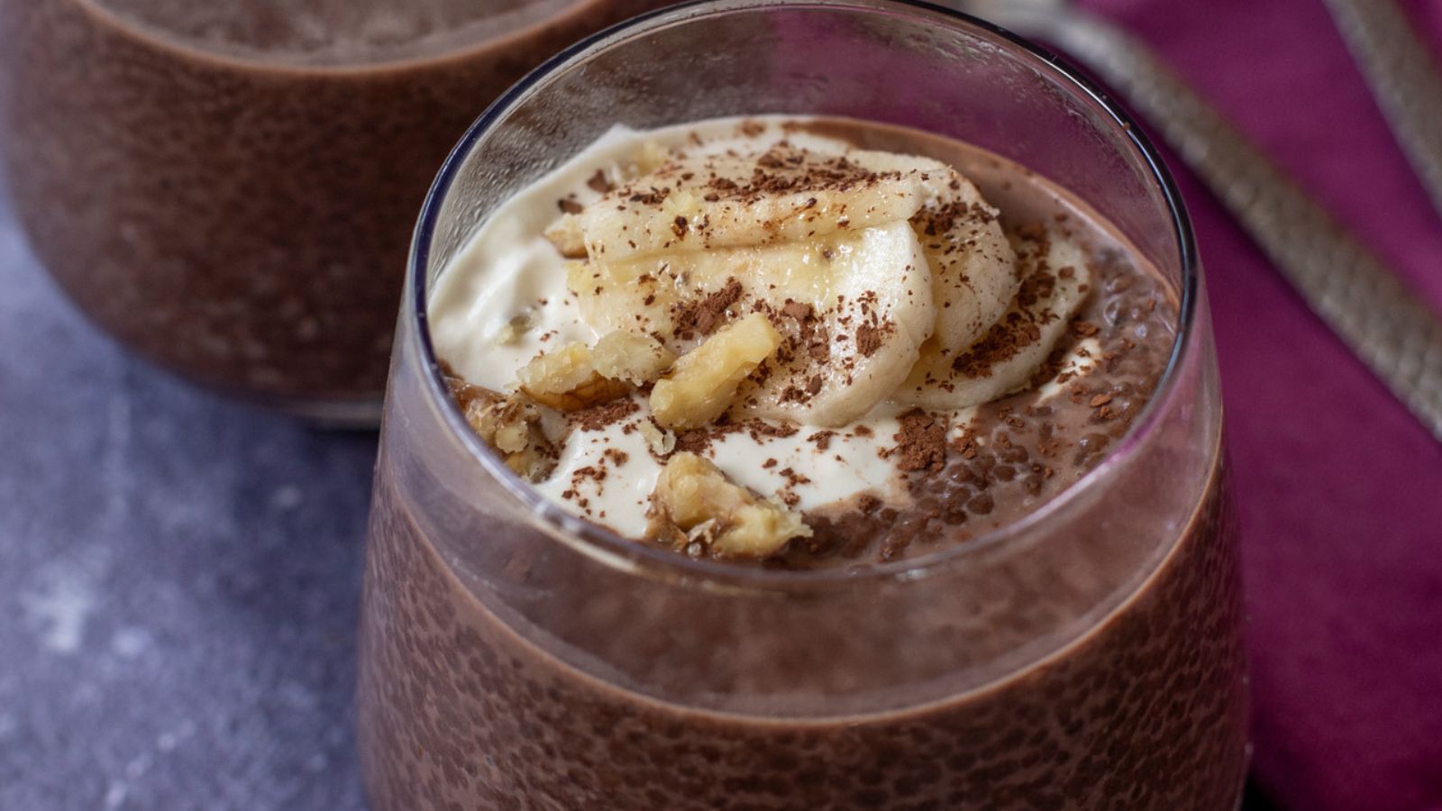 A glass of chocolate chia pudding topped with banana slices, yogurt, chopped walnuts, and chocolate shavings, with another glass partially visible behind it.