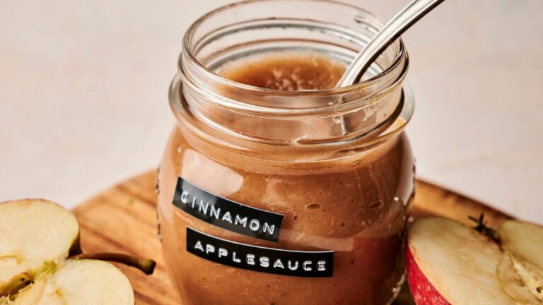 Jar of cinnamon applesauce with a spoon inside, labeled "Cinnamon Applesauce," on a wooden surface, next to sliced apples.