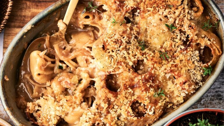 A baked pasta casserole with a golden breadcrumb topping, partially scooped out to reveal creamy pasta shells and garnished with fresh herbs.