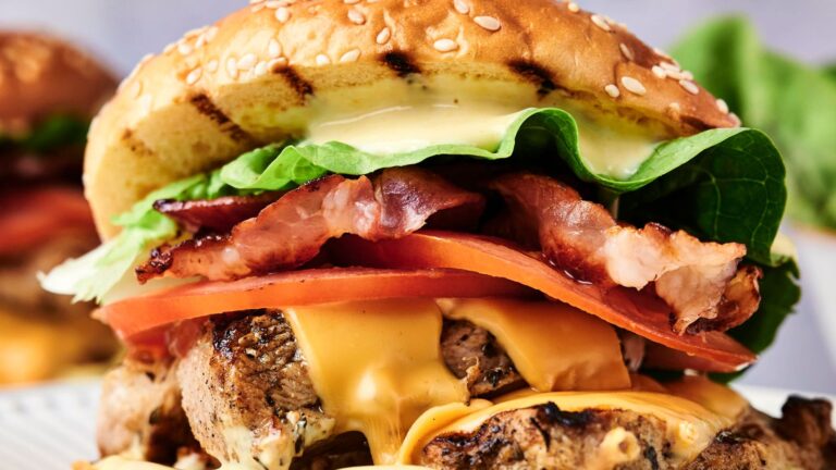 Close-up of a grilled cheeseburger with bacon, lettuce, tomato, and sauce, sandwiched in a sesame seed bun.