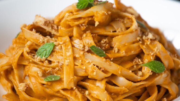 A close-up of wide, flat noodles coated in an orange sauce, garnished with crushed nuts and fresh mint leaves on a white plate.