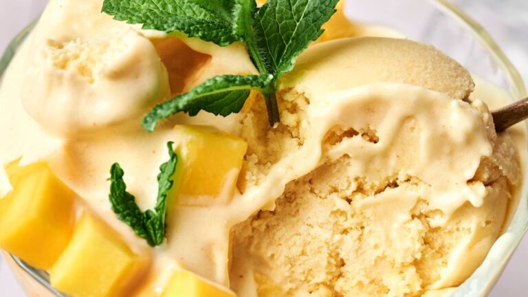 A close-up of a bowl of mango ice cream topped with fresh mango cubes and a sprig of mint.