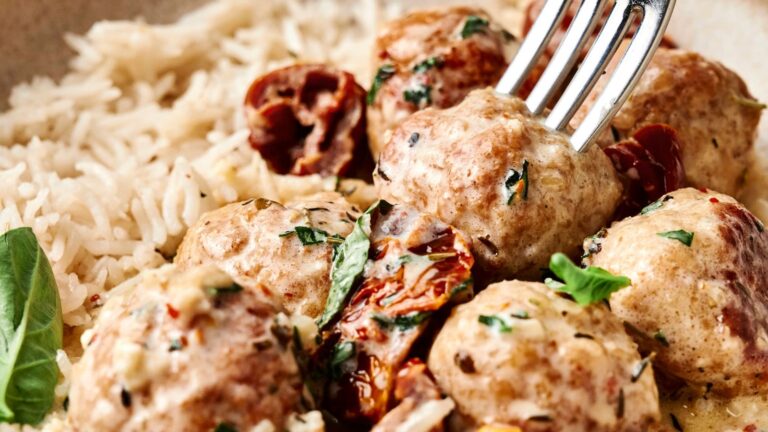 Meatballs covered in a creamy sauce, garnished with herbs, served with white rice and sun-dried tomatoes. A fork is piercing one meatball.