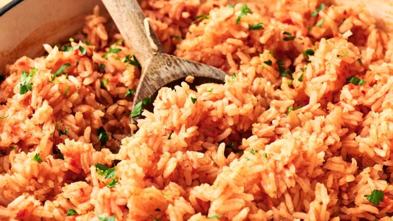 A close-up of cooked Mexican rice in a pan, mixed with tomato sauce and garnished with chopped parsley. A wooden spoon is placed in the rice.