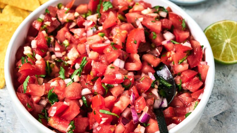 A bowl of freshly made pico de gallo with diced tomatoes, onions, cilantro, and jalapeños, with a spoon inside and a lime wedge nearby.