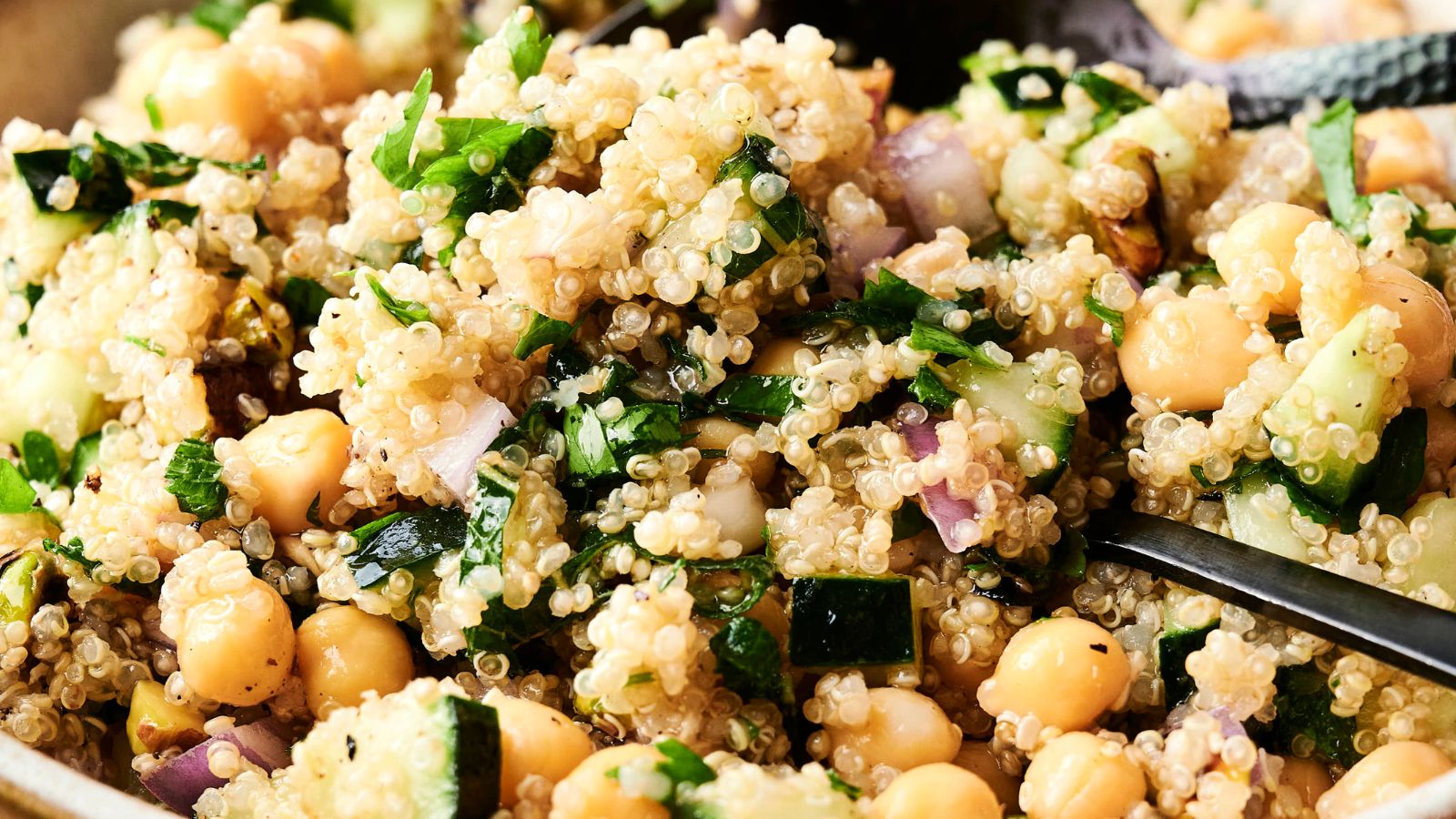 Close-up of a quinoa salad with chickpeas, chopped cucumber, parsley, red onion, and a light dressing, with a spoon partially visible.