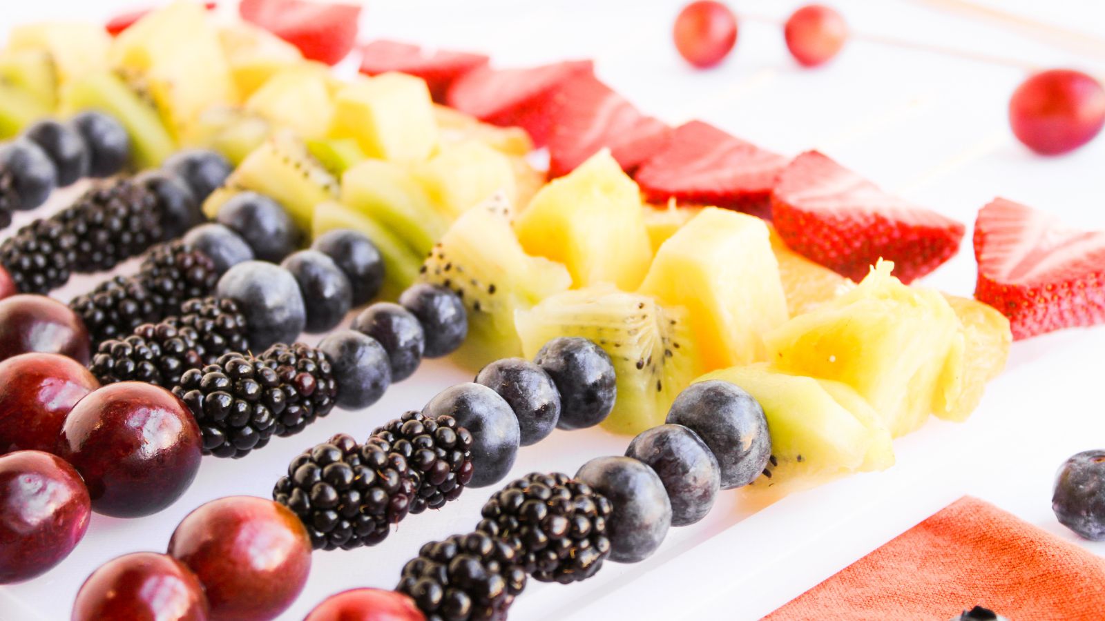 Skewers with grapes, blackberries, blueberries, kiwi, pineapple, and strawberries arranged in neat rows on a white surface.