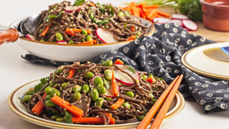 Plates of soba noodle salad with edamame, carrots, radishes, and sesame seeds, accompanied by chopsticks on a patterned cloth.
