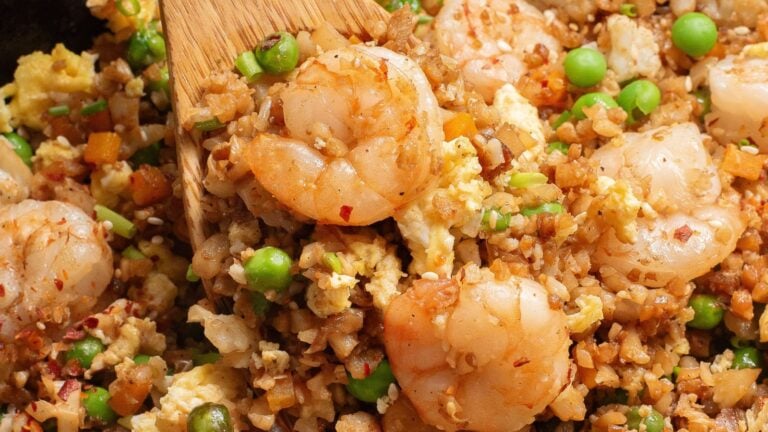 A close-up of shrimp fried rice with peas, scrambled eggs, and diced vegetables being stirred with a wooden spatula.