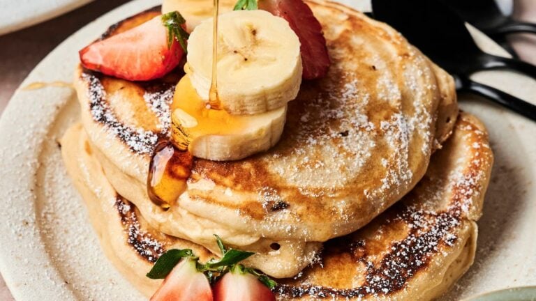 A stack of pancakes topped with sliced bananas, strawberries, and syrup on a plate, with a fork and knife beside it.