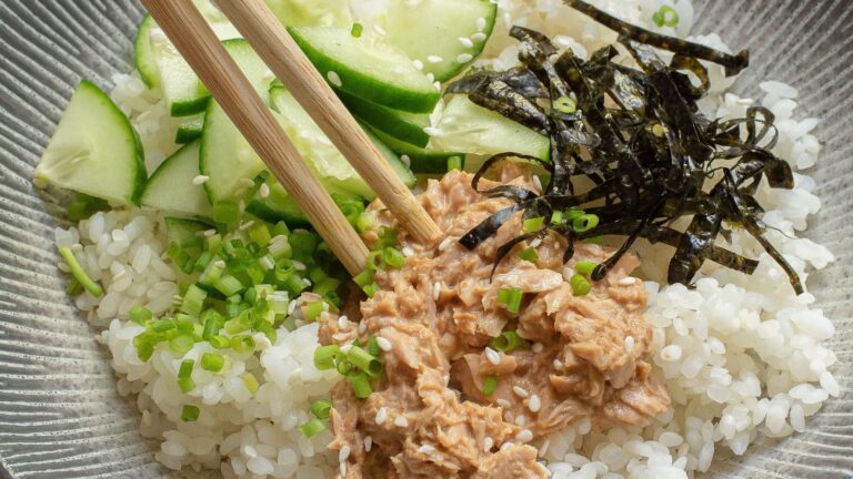 A bowl of white rice topped with chopped cucumber, shredded seaweed, green onions, tuna, and sesame seeds, with wooden chopsticks resting on top.