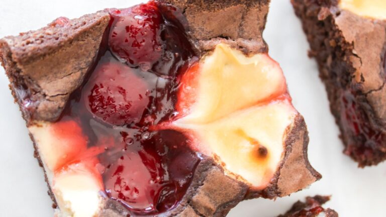A close-up of a brownie square topped with a swirl of cheesecake and cherry filling.