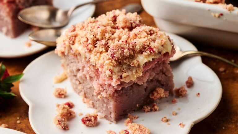 A square slice of crumb-topped strawberry coffee cake sits on a white plate with a spoon beside it.