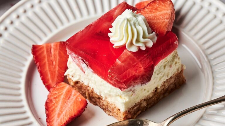A dessert with a biscuit base, creamy layer, and red jelly topped with whipped cream and sliced strawberries on a white plate.