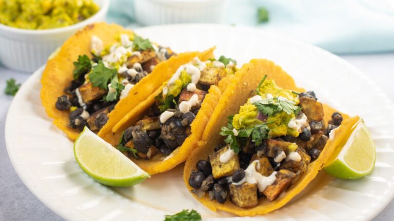 Three tacos filled with black beans, diced vegetables, fresh cilantro, and a creamy sauce are served on a white plate with lime wedges.