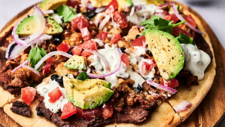 A close-up of a loaded flatbread pizza topped with avocado slices, diced tomatoes, onion, cilantro, seasoned meat, black beans, and a drizzle of creamy sauce.