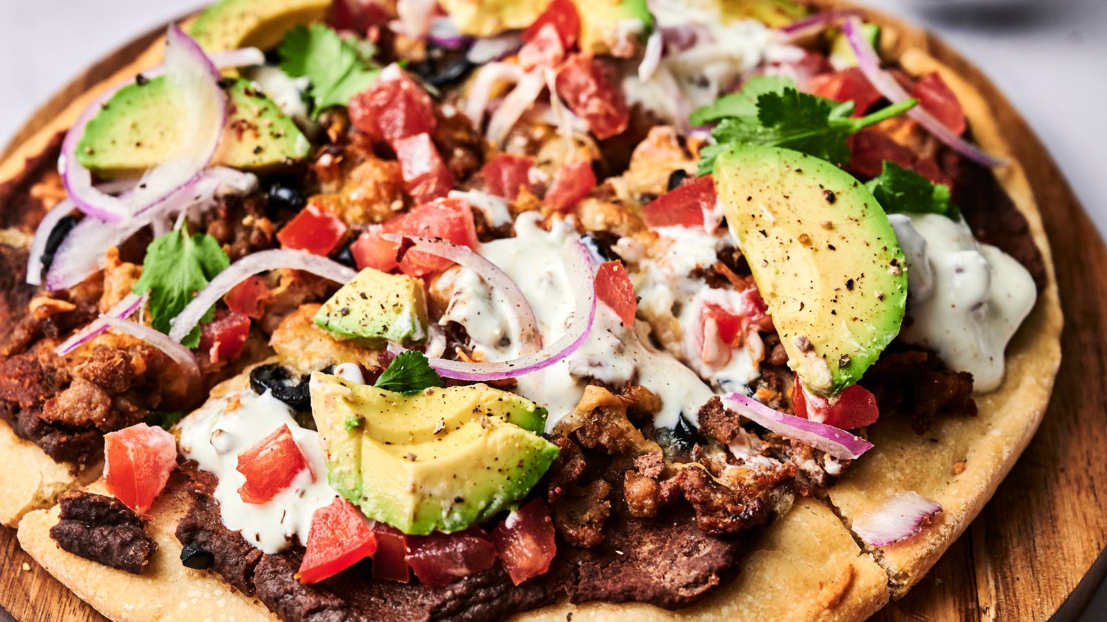 A close-up of a loaded flatbread pizza topped with avocado slices, diced tomatoes, onion, cilantro, seasoned meat, black beans, and a drizzle of creamy sauce.