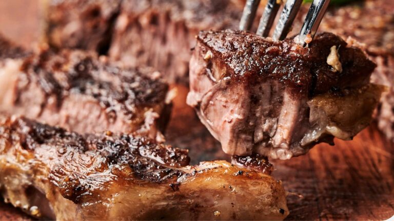 Close-up of a grilled steak cube being lifted by a fork. The meat is juicy with visible char marks and marbling, set on a wooden surface with additional steak pieces in the background.