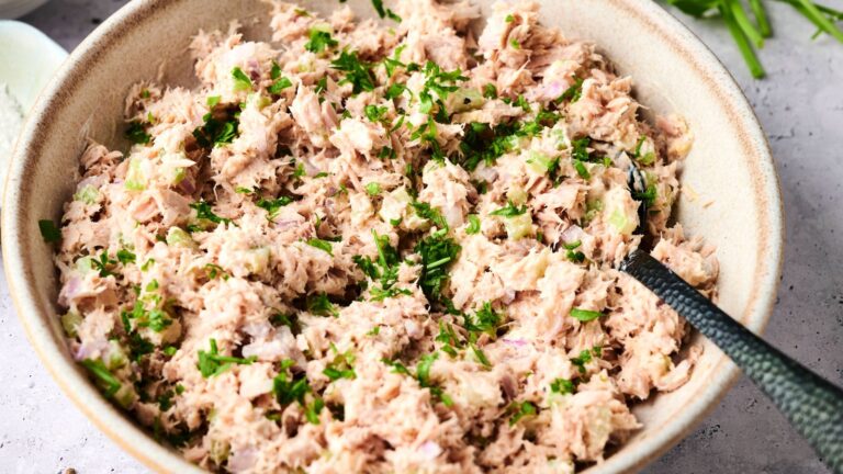 A bowl filled with tuna salad mixed with herbs and finely chopped onions, with a spoon resting inside.