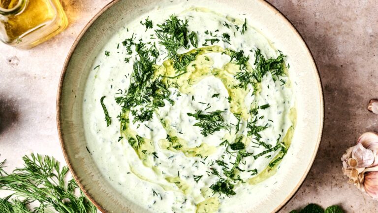A bowl of creamy tzatziki garnished with chopped dill and a drizzle of olive oil. Fresh herbs and a garlic bulb are visible nearby.