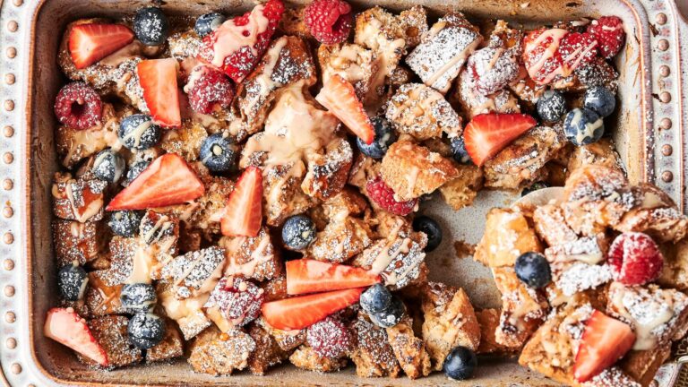 Baked French toast casserole topped with strawberries, blueberries, raspberries, powdered sugar, and a drizzle of sauce in a metal baking dish.