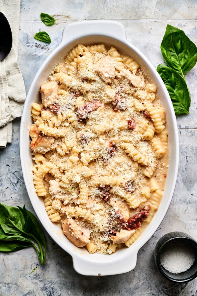A white dish with baked pasta, chunks of chicken, sun-dried tomatoes, and breadcrumbs resembles a Marry Me Chicken Casserole, garnished with fresh basil leaves on the side.