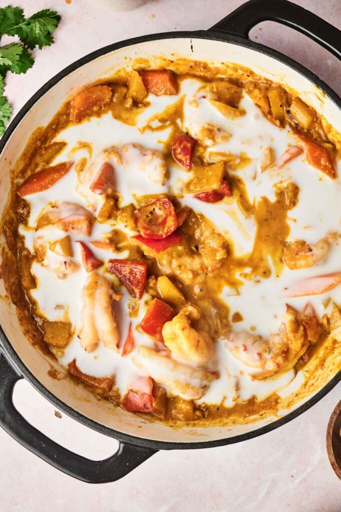 A pot of Massaman prawn curry with chunks of vegetables and coconut milk, garnished with cilantro, in a black-handled pan on a light surface.