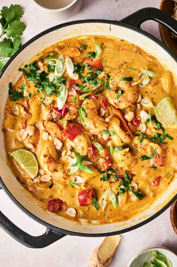 A creamy Massaman curry stew in a pot, featuring shrimp, chopped vegetables, lime wedges, peanuts, and fresh herbs.