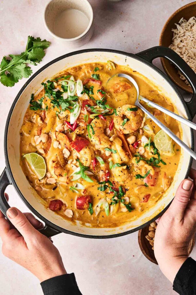 A person holds a pot of creamy Massaman curry with sliced lime, peanuts, red bell peppers, and greens. A serving spoon rests in the dish. Rice and garnishes are nearby on a light-colored surface.