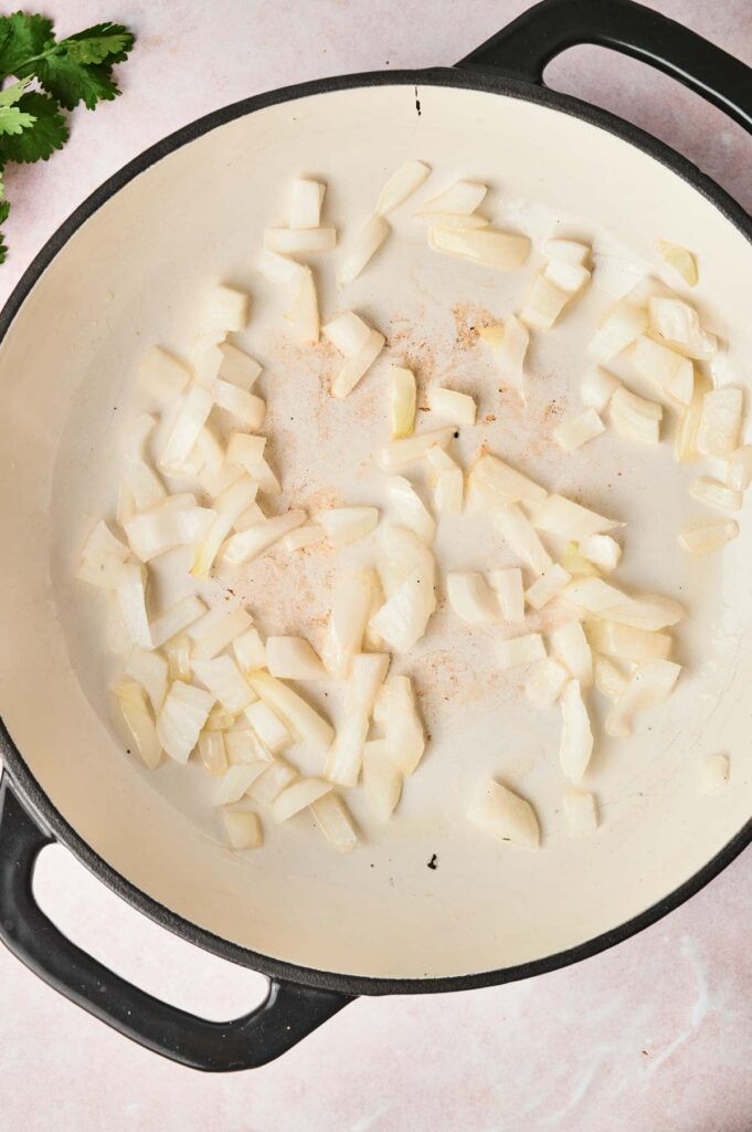 Chopped onions are gently saut&eacute;ing in a large black skillet, creating the perfect base for a rich Massaman Curry on a light-colored surface.