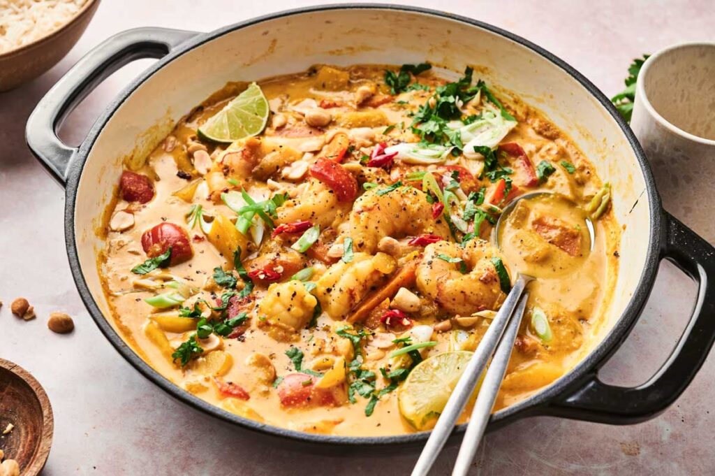A pot of creamy Massaman curry with shrimp, garnished with green onions, herbs, and lime slices.