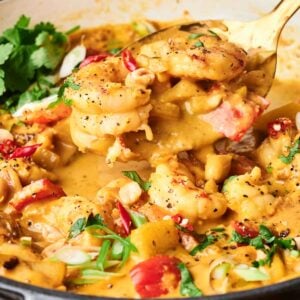 A skillet filled with creamy shrimp massaman curry, featuring shrimp, diced red chili, herbs, and green onions, with a serving spoon lifting some out.
