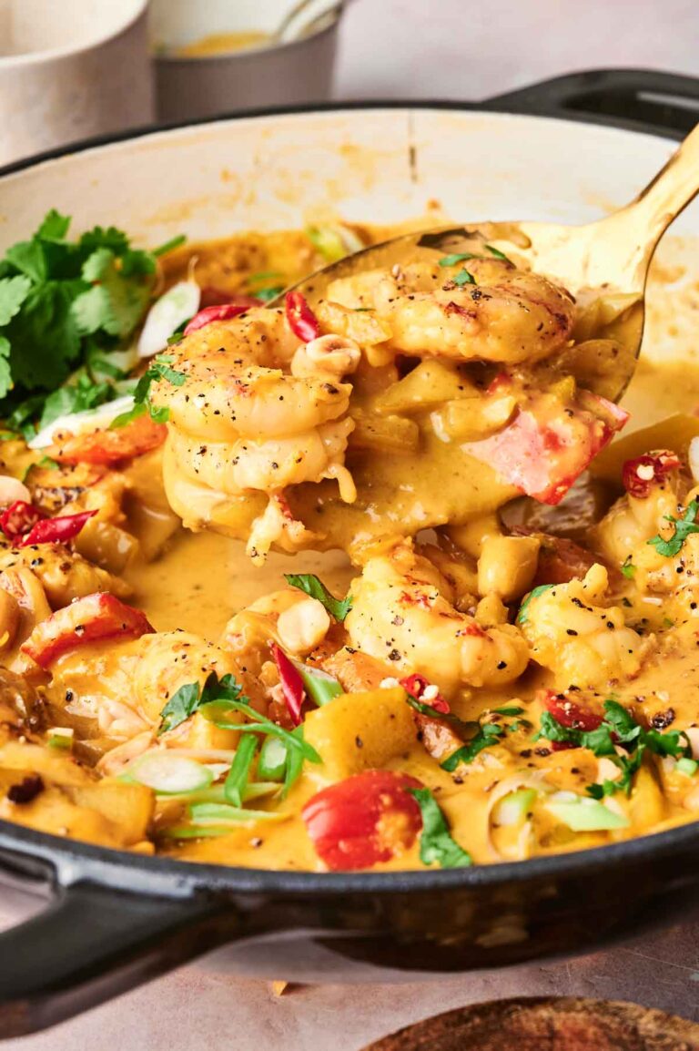A skillet filled with creamy shrimp massaman curry, featuring shrimp, diced red chili, herbs, and green onions, with a serving spoon lifting some out.