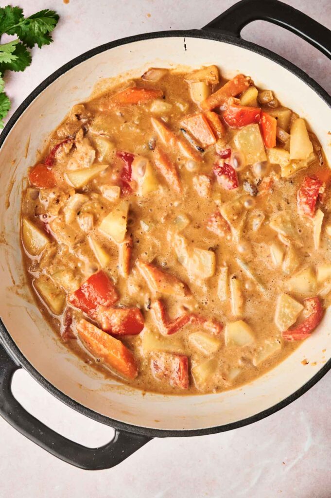 A pot of creamy Massaman Curry stew with chunks of potatoes, carrots, and peppers in a thick sauce. The pot has black handles and is set against a light background.