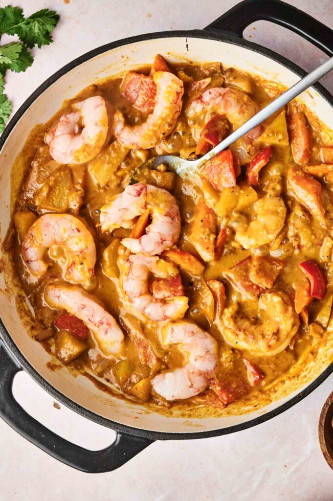 A pan holds a Massaman Curry dish with shrimp and sliced vegetables, including red peppers. A spoon rests in the creamy delight, with a sprig of cilantro adding a fresh touch on the side.