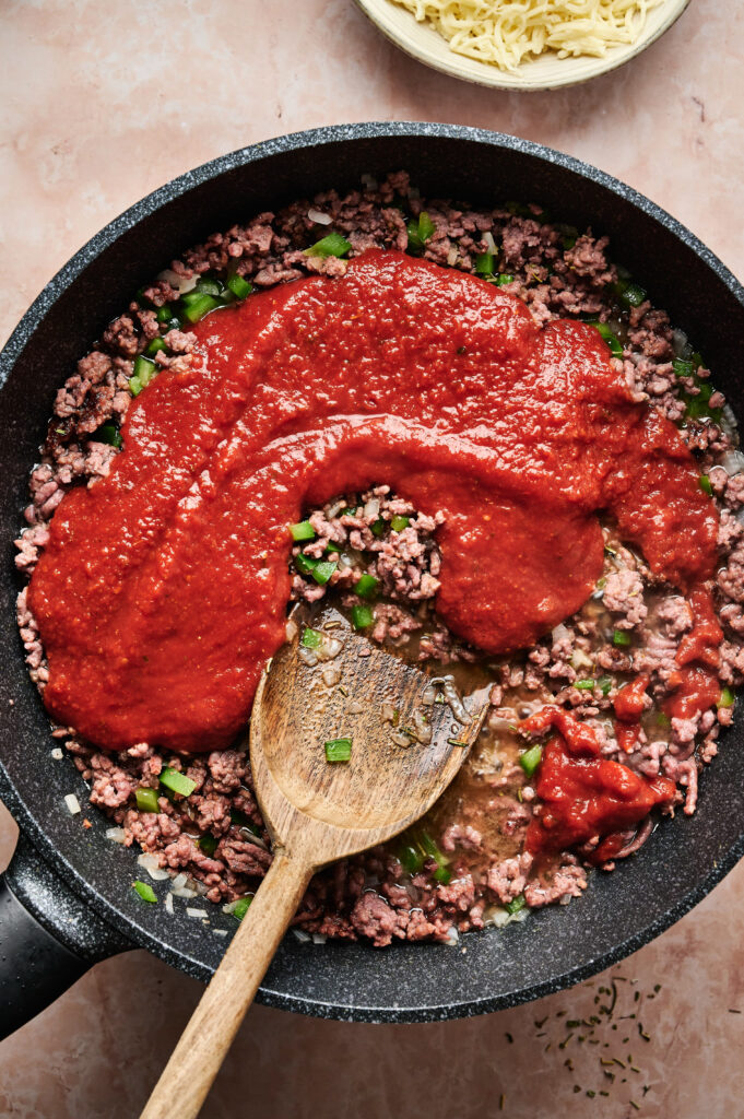A black pan contains cooking ground meat and chopped green peppers, resembling a pizza casserole, with a wooden spoon amidst a generous topping of red tomato sauce. A bowl of shredded cheese tempts in the background.