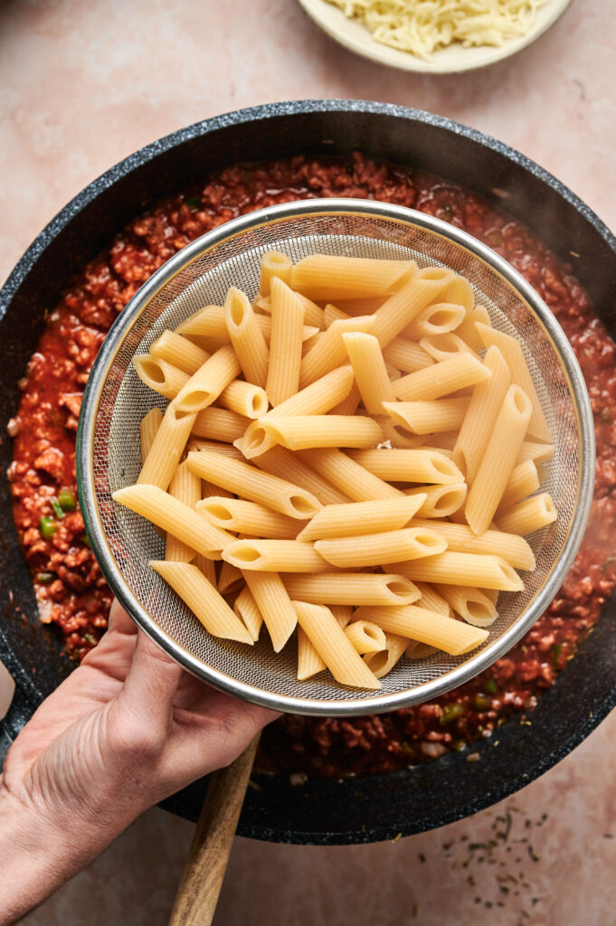 A hand holds a sieve with cooked penne pasta over a pan of rich tomato and meat sauce, ready for mixing into a pizza casserole delight.
