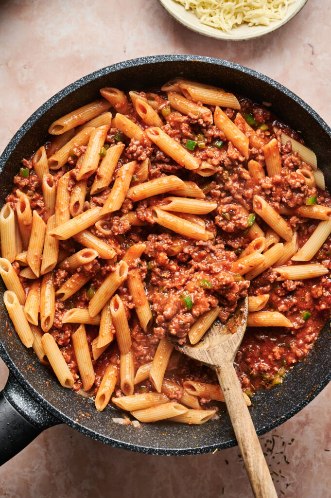 A pan filled with cooked penne pasta, reminiscent of a pizza casserole, is mixed with ground meat and tomato sauce. A wooden spoon rests in the pan.