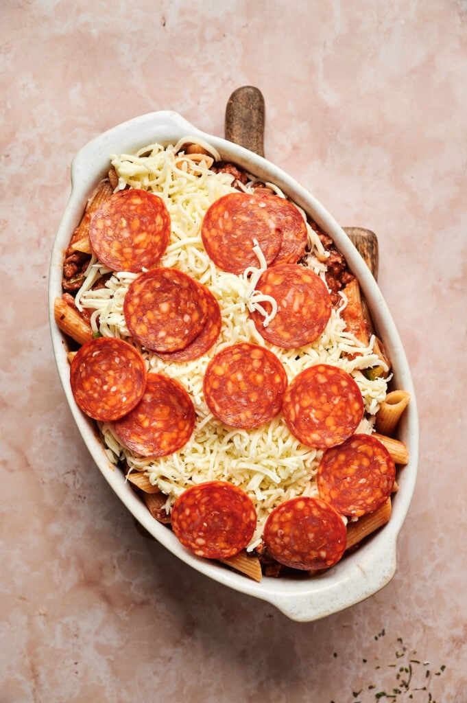An oval baking dish brimming with pizza casserole, topped with uncooked pasta layered beneath melted cheese, pepperoni slices, and rich sauce rests on a textured surface.