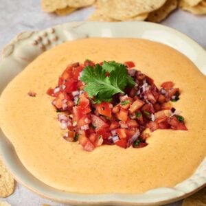 A bowl of creamy queso dip adorned with diced tomato salsa nestles in the center, garnished with cilantro. Tortilla chips are scattered around the bowl on a light surface.
