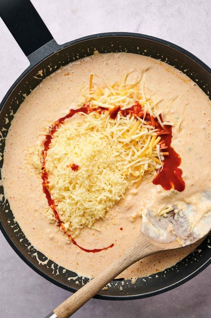 Queso dip and red sauce meld into the creamy mixture in a pan, with a wooden spoon resting on the side.