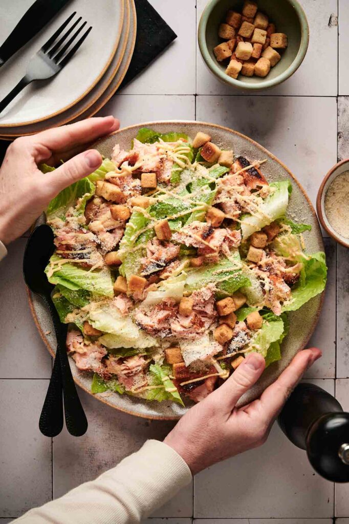 Hands expertly preparing a salmon Caesar salad with croutons and grated cheese on a table, next to utensils and a small bowl of croutons.