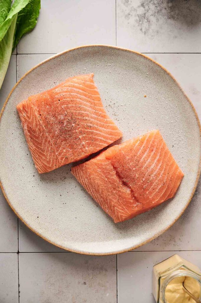 Two raw salmon fillets, perfect for a fresh salmon Caesar salad, are sprinkled with seasoning on a round, light-colored plate. Nearby, a crisp lettuce leaf rests on the tiled surface.