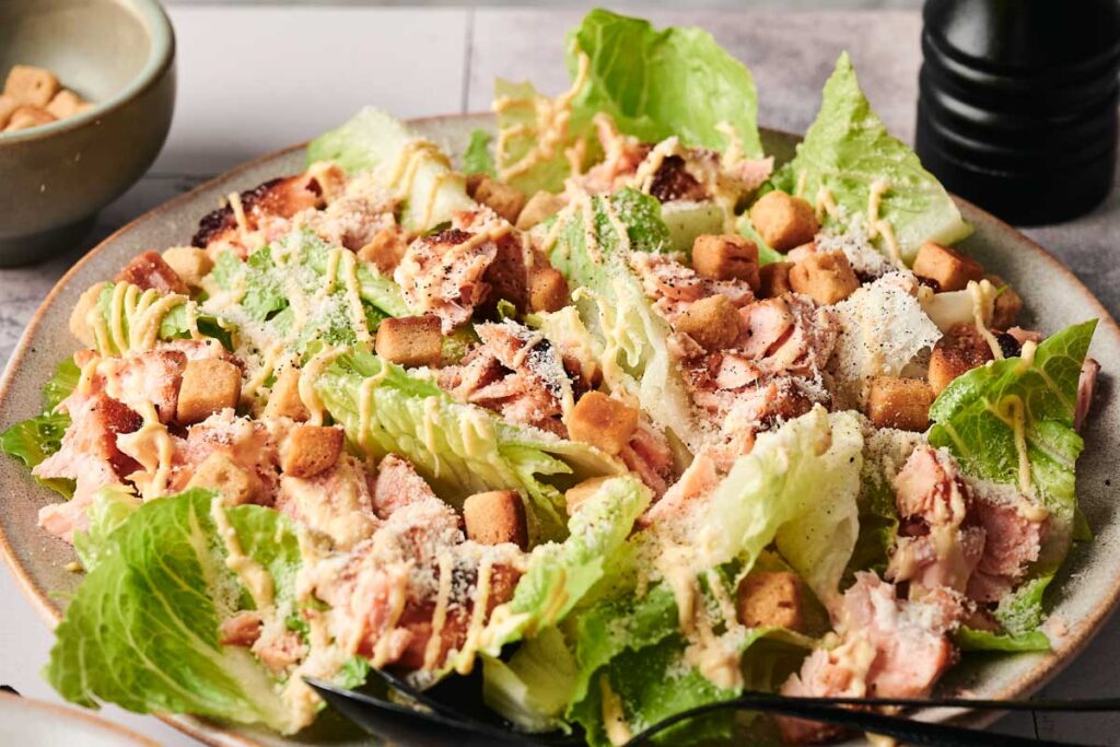A salmon Caesar salad featuring romaine lettuce, croutons, and grated cheese is artfully arranged on a plate. In the background, a bowl of extra croutons and a pepper mill complete this culinary masterpiece.