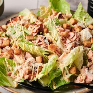 A salmon Caesar salad featuring romaine lettuce, croutons, and shredded cheese, all drizzled with dressing on a plate.