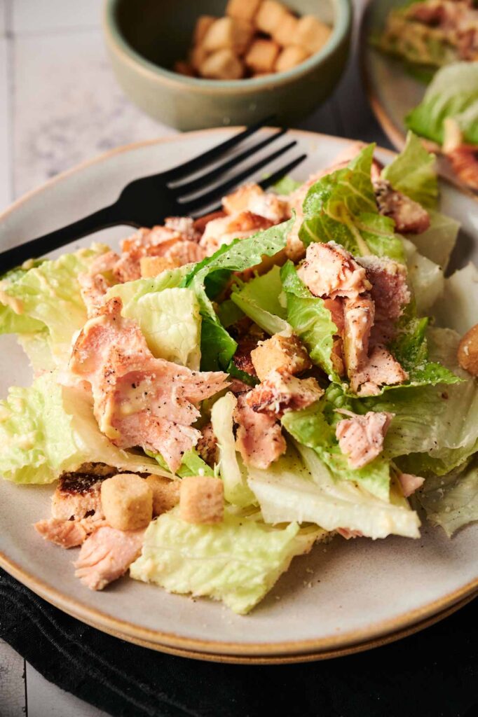 A plate of salmon Caesar salad with lettuce, tender salmon pieces, and crispy croutons sits invitingly beside a sleek black fork. In the background, a bowl brimming with extra croutons tempts you to indulge further.
