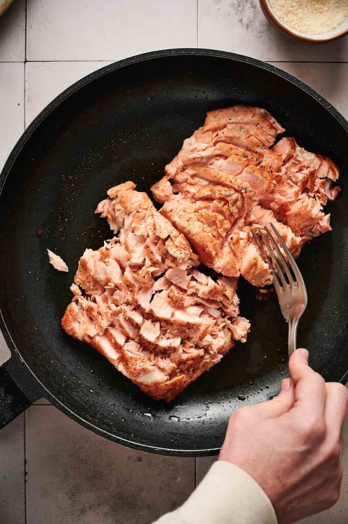 A person flaking cooked salmon with a fork in a black frying pan on a tiled countertop, preparing it for a delicious salmon Caesar salad.
